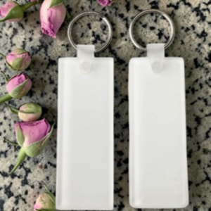 Two blank white rectangular keychain tags with silver rings on a speckled countertop, beside pink rose buds.