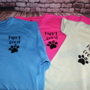 Three colorful t-shirts (blue, pink, and cream) laid out on a brick surface, each with 'Puppy crew' and a paw print graphic.