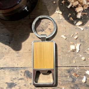 Wood-paneled bottle opener keychain on a rustic wooden surface with wood shavings nearby.