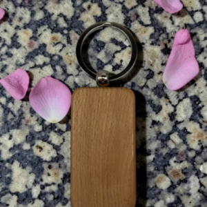 Wooden rectangular keychain with a metal ring on a speckled carpet, pink flower petals around it.