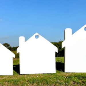 Outdoor art installation of oversized white house silhouettes standing on a grassy field under a blue sky