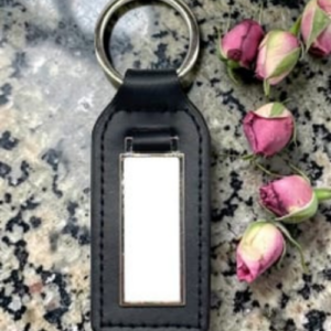Leather keychain with a blank rectangular metal tag, placed on a speckled surface beside pink rose buds.