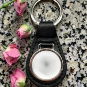 Keychain with a round silver metal case set in a black leather holder on a speckled countertop, surrounded by pink rosebuds.