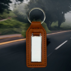 Brown leather keychain with a silver rectangular metal tag, set against a blurred roadway background.