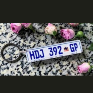 Blue-on-white license plate keychain reading 'HDJ 392 GP' lying on a speckled countertop with pink roses nearby.