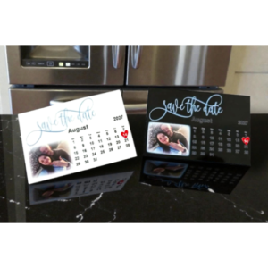 Two desk calendar cards reading 'Save the Date' in a kitchen, one white and one black, showing August 2027 and a small couple photo, placed on a dark countertop.